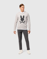 A man stands against a plain background, exuding timeless style in a light gray Psycho Bunny MENS LINCOLN MERINO WOOL SWEATER - B6E831D200 featuring a black bunny and crossbones design, layered over a white shirt, paired with dark gray pants and white sneakers.