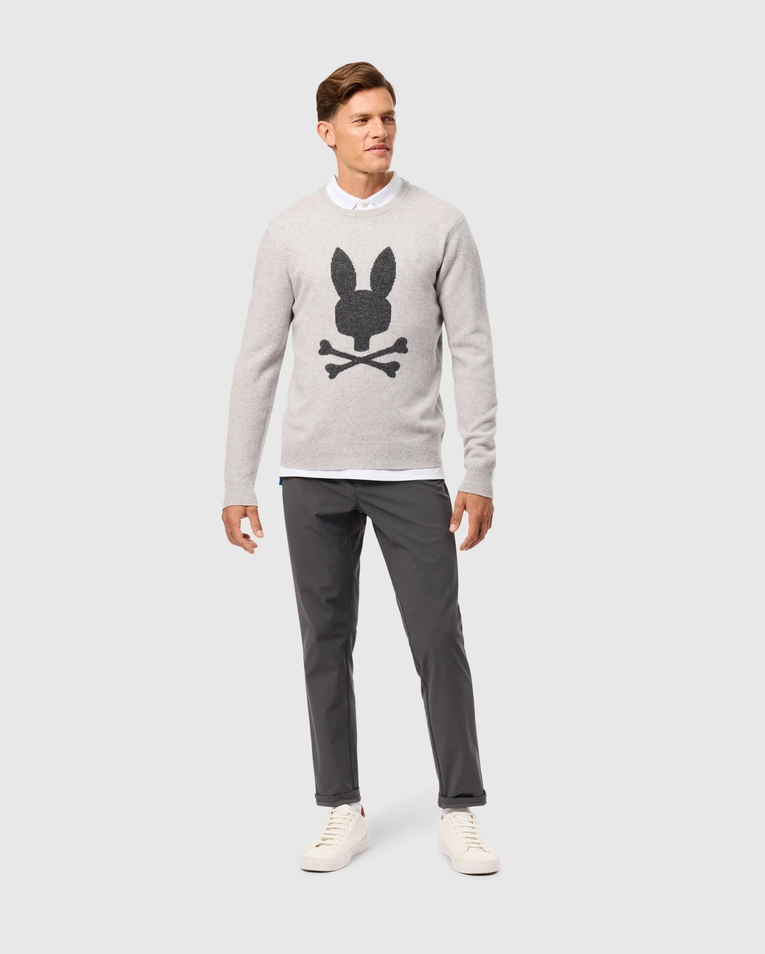 A man stands against a plain background, exuding timeless style in a light gray Psycho Bunny MENS LINCOLN MERINO WOOL SWEATER - B6E831D200 featuring a black bunny and crossbones design, layered over a white shirt, paired with dark gray pants and white sneakers.