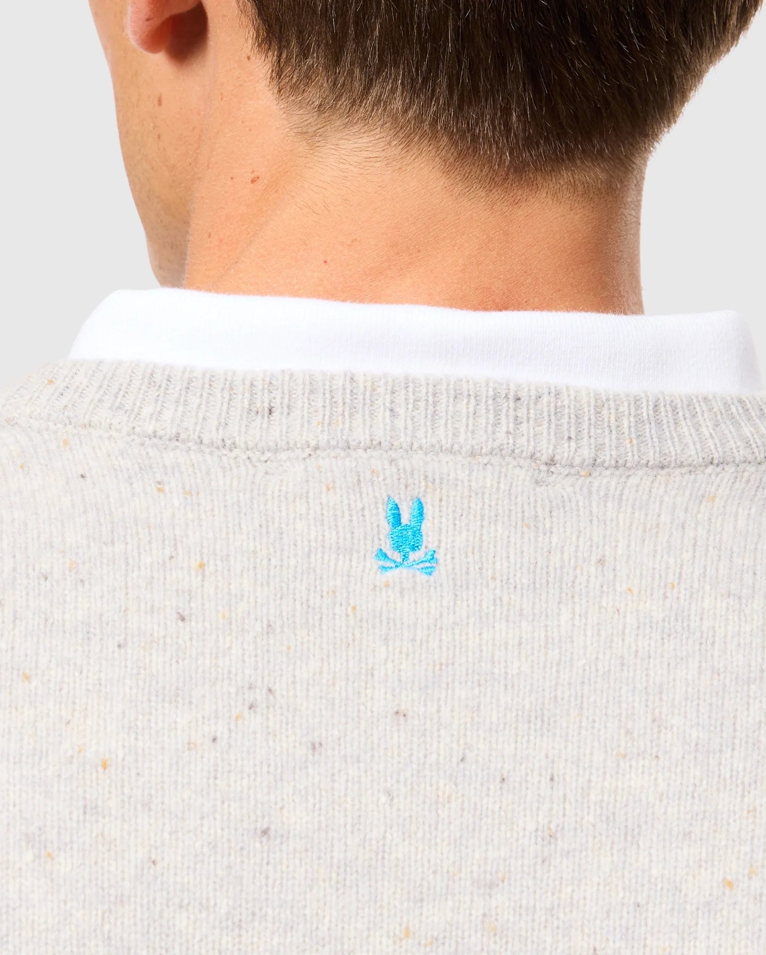 Close-up of the back of a person's neck adorned in the Psycho Bunny Men's Lincoln Merino Wool Sweater (B6E831D200), crafted from soft merino wool. This light gray piece with multicolored specks features a timeless style, showcasing the iconic blue embroidered bunny logo just below the white collar of the shirt underneath.