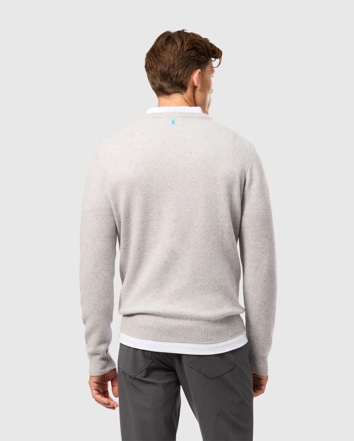 A person with short brown hair is standing against a plain background, facing away and wearing the Psycho Bunny light gray MENS LINCOLN MERINO WOOL SWEATER (B6E831D200). The sweater, showcasing timeless style, is paired with dark pants and has a subtle blue logo near the neckline.