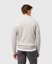 A person with short brown hair is standing against a plain background, facing away and wearing the Psycho Bunny light gray MENS LINCOLN MERINO WOOL SWEATER (B6E831D200). The sweater, showcasing timeless style, is paired with dark pants and has a subtle blue logo near the neckline.