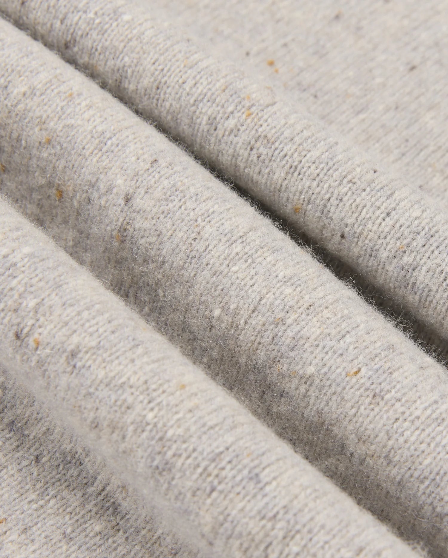 A close-up of the MENS LINCOLN MERINO WOOL SWEATER - B6E831D200 by Psycho Bunny highlights its textured merino wool fabric in light gray, adorned with subtle speckles of brown and beige. This soft, slightly ribbed surface creates gentle folds and shadows, showcasing a timeless style.