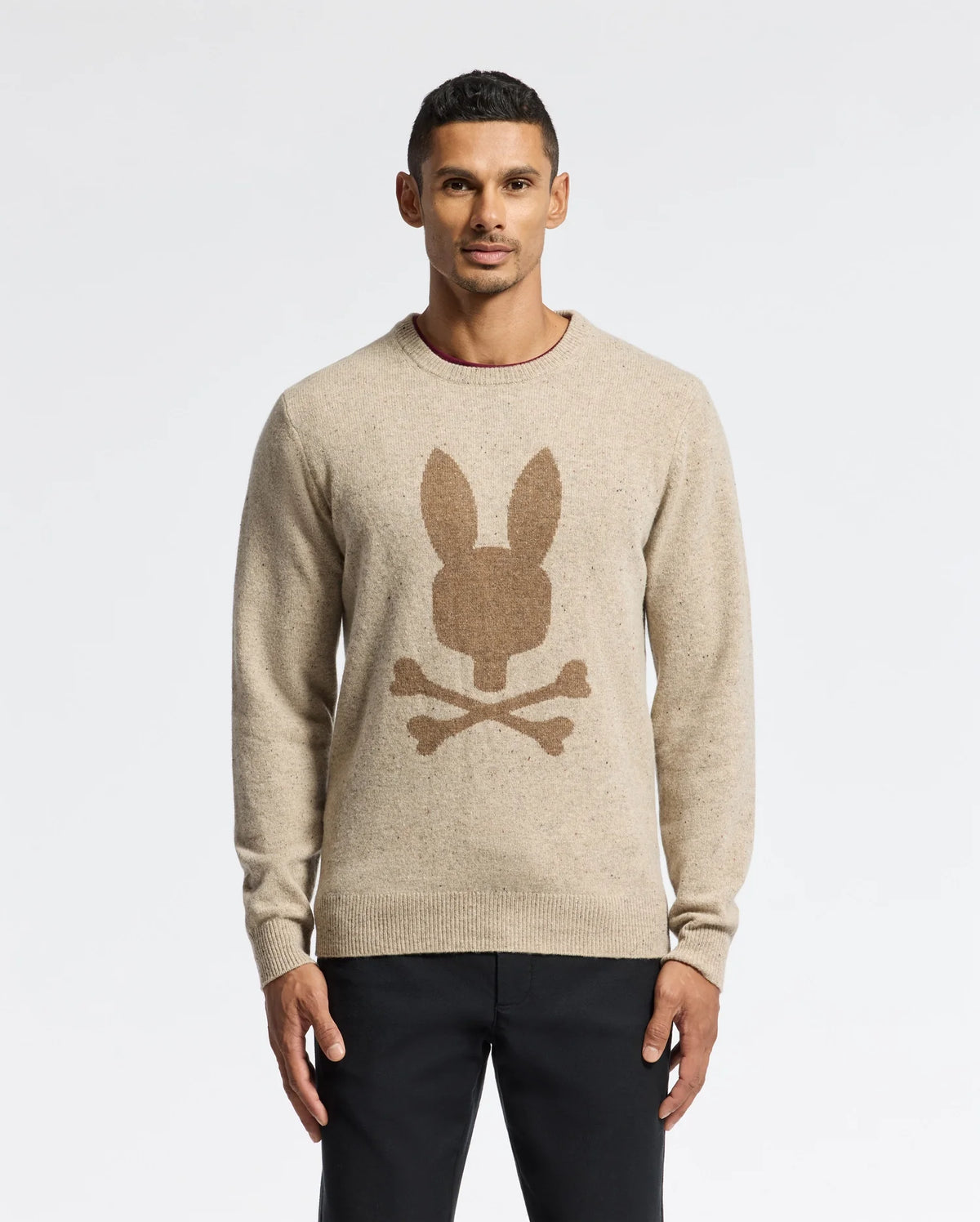 A person is wearing the Psycho Bunny Men's Lincoln Merino Wool Sweater (B6E831E200) in beige, which showcases a bunny head and crossbones design on the front. They are standing against a plain white background, dressed in black pants and looking at the camera.