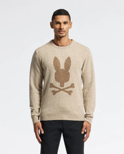 A person is wearing the Psycho Bunny Men's Lincoln Merino Wool Sweater (B6E831E200) in beige, which showcases a bunny head and crossbones design on the front. They are standing against a plain white background, dressed in black pants and looking at the camera.