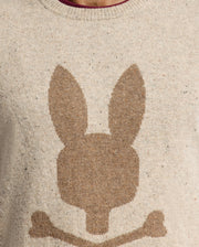 The MENS LINCOLN MERINO WOOL SWEATER - B6E831E200 by Psycho Bunny is showcased in a close-up image. This beige sweater features a brown silhouette of a stylized rabbit head with long ears above two crossed bones, highlighting the texture of the warm Merino wool knit fabric.