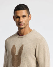A person with short dark hair is wearing the Psycho Bunny Men's Lincoln Merino Wool Sweater (B6E831E200), a beige sweater featuring a warm merino wool construction and adorned with a brown silhouette of rabbit ears on the front. The backdrop is plain white.