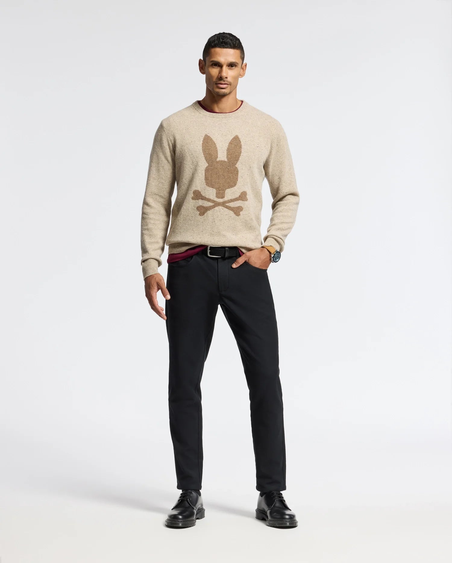 A man stands confidently against a plain background, hands in his pockets, wearing the warm MENS LINCOLN MERINO WOOL SWEATER by Psycho Bunny, adorned with a distinctive bunny and crossbones design. Paired with black pants and shoes, he exudes a relaxed style perfect for any occasion.