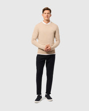 A man stands against a plain background wearing a beige Psycho Bunny MENS BELMORE EXTRA FINE COTTON SWEATER - B6E832D200 over a white shirt, black pants, and dark sneakers. The sweater features premium stitching with an embroidered tonal Bunny logo. His hands are clasped in front as he looks forward.