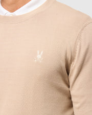 A close-up of a person wearing the Psycho Bunny MENS BELMORE EXTRA FINE COTTON SWEATER (B6E832D200) with an embroidered Bunny logo and crossbones on the chest, layered over a white shirt whose collar peeks from underneath.