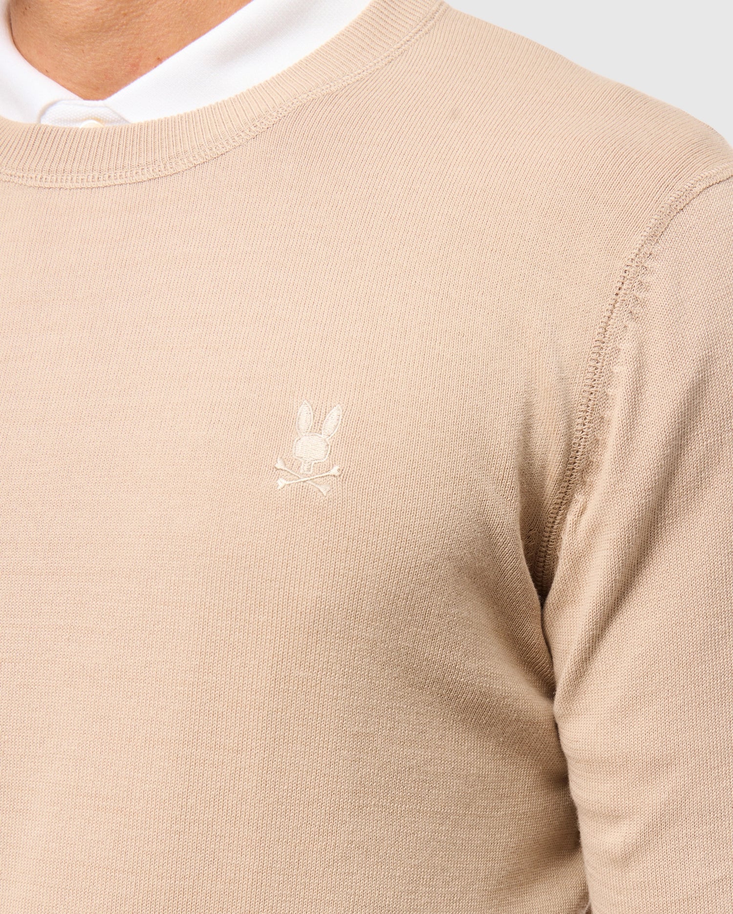 A close-up of a person wearing the Psycho Bunny MENS BELMORE EXTRA FINE COTTON SWEATER (B6E832D200) with an embroidered Bunny logo and crossbones on the chest, layered over a white shirt whose collar peeks from underneath.