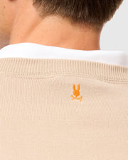 A close-up shows a person's neck wearing the Psycho Bunny's MENS BELMORE EXTRA FINE COTTON SWEATER - B6E832D200, featuring an orange Bunny logo with crossed bones on the back. The beige sweater nicely complements a white collar underneath.