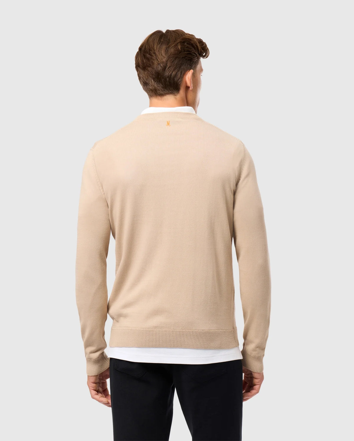 A brown-haired man, seen from behind, wears a beige Psycho Bunny MENS BELMORE EXTRA FINE COTTON SWEATER (B6E832D200) over a white shirt and black pants. The embroidered Bunny logo adds subtlety as he stands against a neutral gray background.