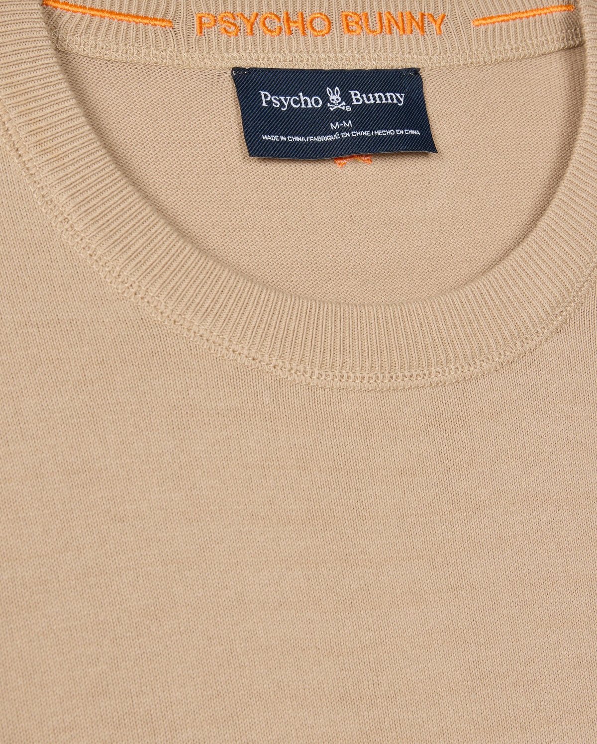 A close-up of the MENS BELMORE EXTRA FINE COTTON SWEATER - B6E832D200 by Psycho Bunny shows its beige fabric, ribbed neckline, and tag with brand details. The inner collar displays "PSYCHO BUNNY" in vibrant orange, and a tonal embroidered Bunny logo adds subtle elegance.