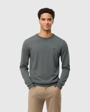 A man with short curly hair stands smiling with his hands in his pockets, wearing the high-end MENS BELMORE COTTON SWEATER by Psycho Bunny. The sweater is intricately embroidered and dark green, paired with beige pants, all set against a simple light gray background.