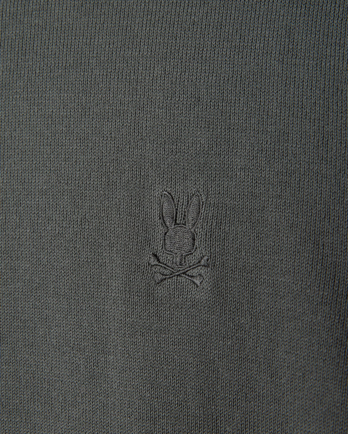 The MENS BELMORE COTTON SWEATER - B6E832D200 by Psycho Bunny features a detailed close-up of its luxurious dark green textured cotton fabric, highlighted with the brand's signature small embroidered bunny head logo above crossed bones, centered prominently on the garment.
