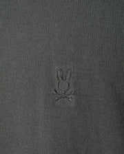 The MENS BELMORE COTTON SWEATER - B6E832D200 by Psycho Bunny features a detailed close-up of its luxurious dark green textured cotton fabric, highlighted with the brand's signature small embroidered bunny head logo above crossed bones, centered prominently on the garment.