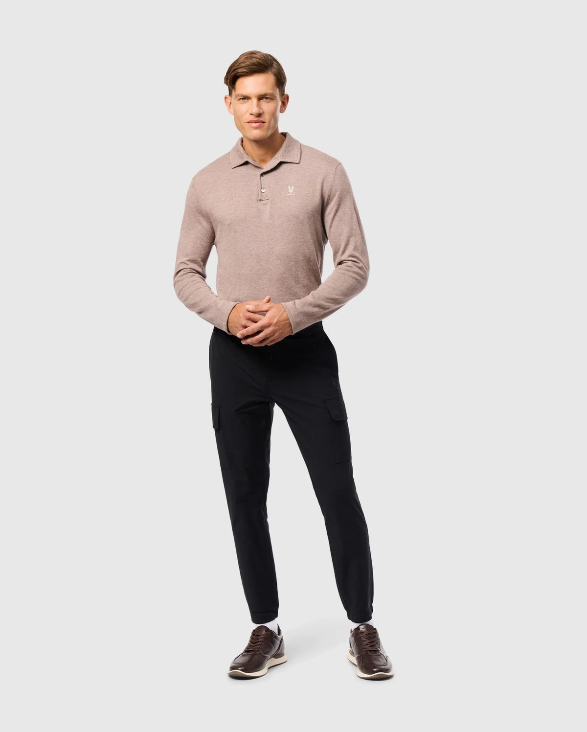 A man stands confidently wearing the luxurious Psycho Bunny MENS BRADSHAW POLO SWEATER (B6E835D200) in light brown, paired with black pants and dark brown shoes. He has short brown hair and poses against a plain gray background.