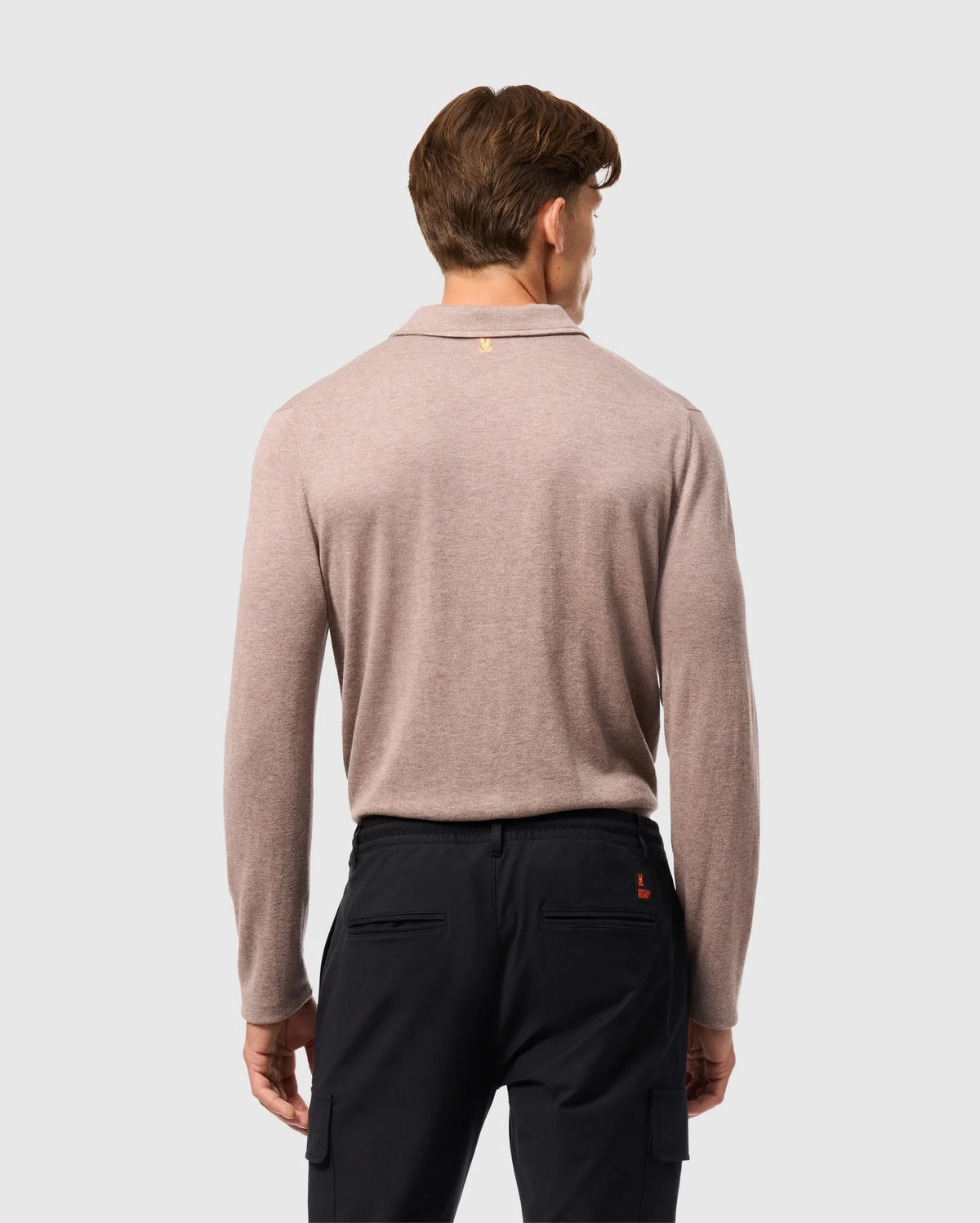 A person with short brown hair stands back to the camera, wearing a Psycho Bunny MENS BRADSHAW POLO SWEATER in light brown paired with black pants. The plain, light gray backdrop accentuates the elegance of this luxurious merino wool attire.