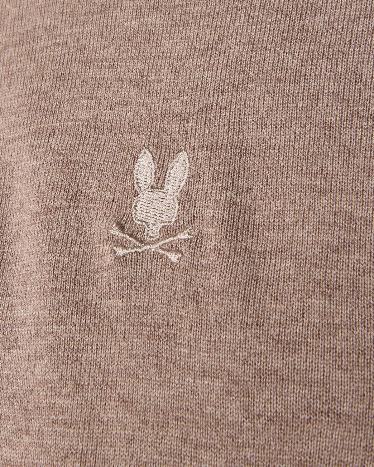 Detailed view of the brown MENS BRADSHAW POLO SWEATER by Psycho Bunny, crafted from luxurious merino wool. It features a small, white embroidered bunny head with crossed bones beneath it, adding a subtle detail to the fine texture of the fabric.