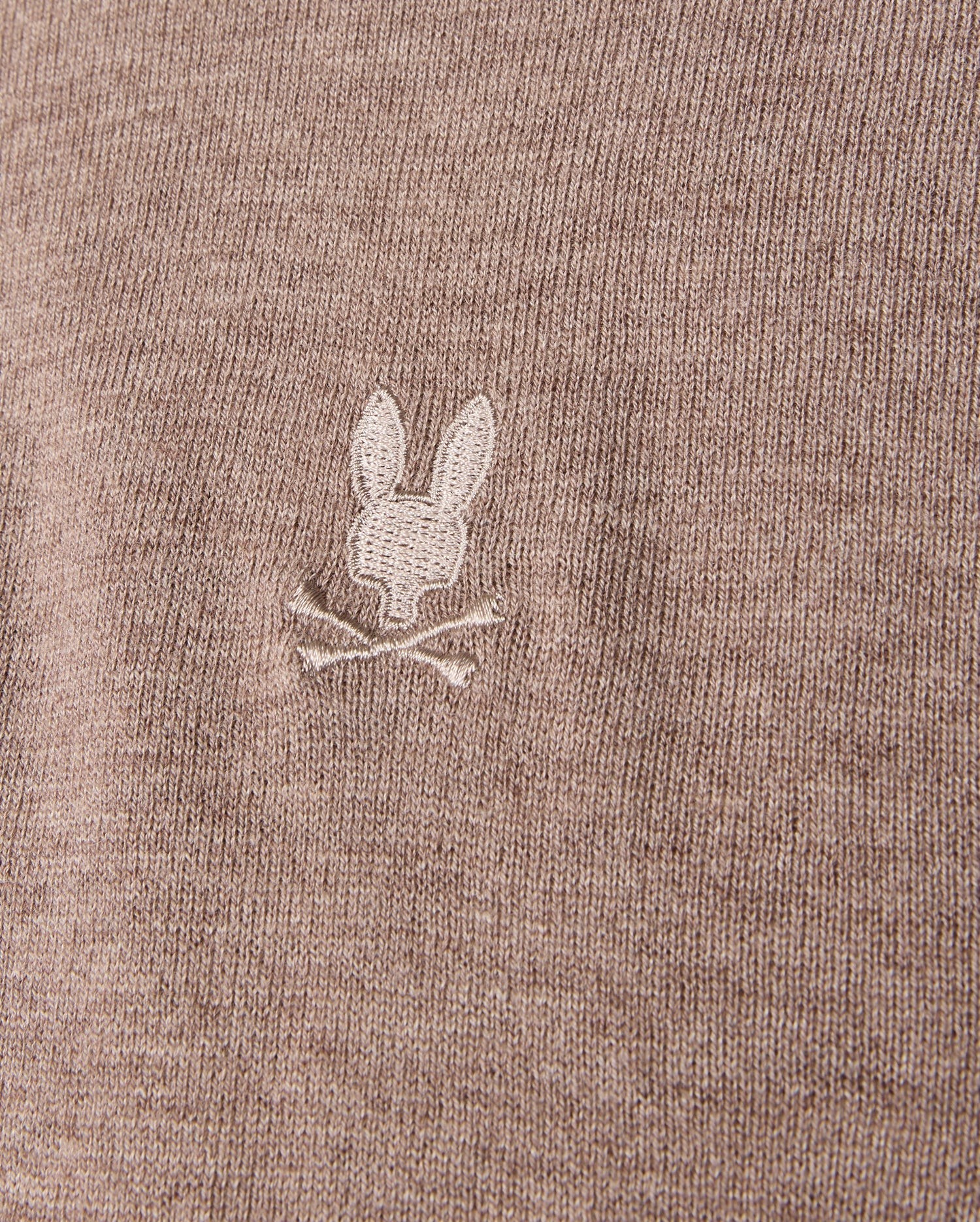 Detailed view of the brown MENS BRADSHAW POLO SWEATER by Psycho Bunny, crafted from luxurious merino wool. It features a small, white embroidered bunny head with crossed bones beneath it, adding a subtle detail to the fine texture of the fabric.
