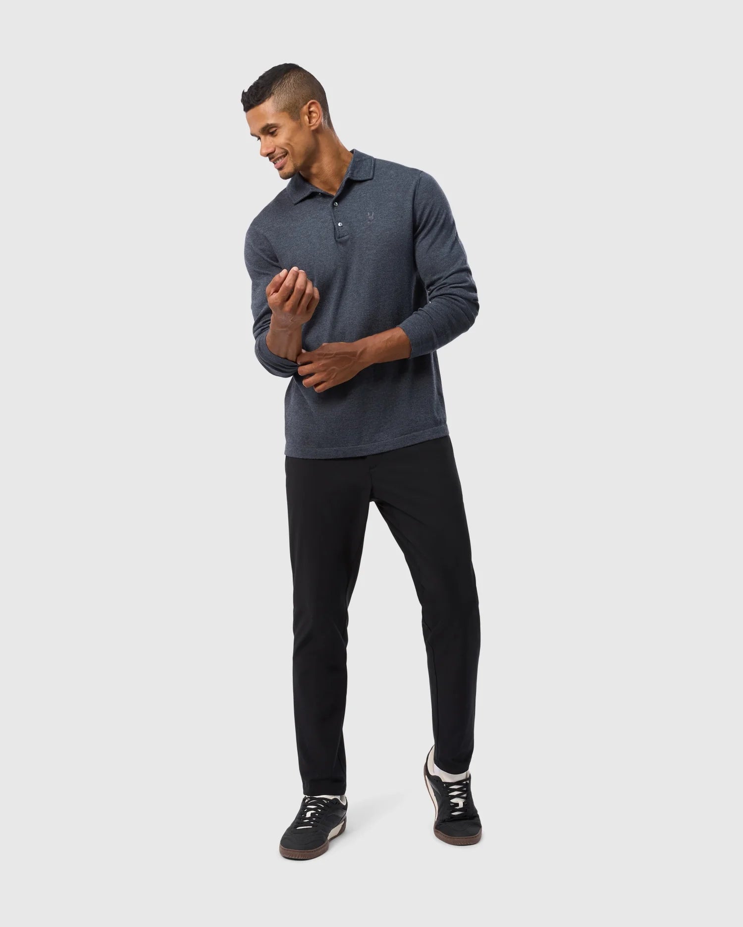 A man in a relaxed pose, wearing the Psycho Bunny MENS BRADSHAW POLO SWEATER - B6E835D200, paired with black pants, adjusts his sleeve while looking down. He has short hair and a subtle smile, standing against a simple light grey background that highlights his comfort and style.