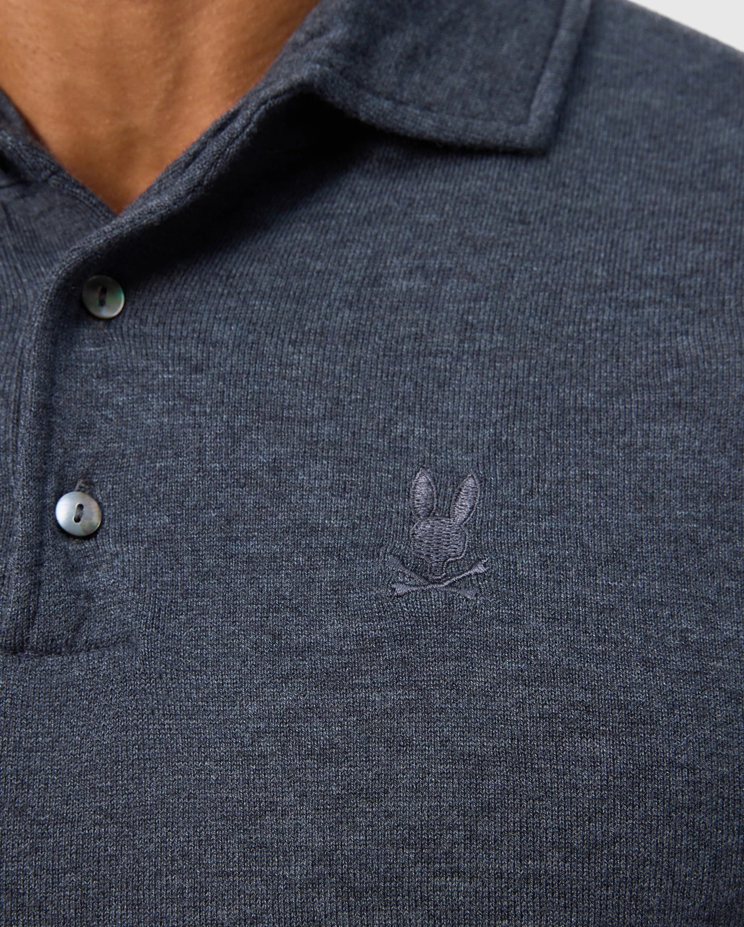 Close-up of the MENS BRADSHAW POLO SWEATER - B6E835D200 by Psycho Bunny, a dark grey merino wool men's polo featuring two buttons and an embroidered logo with a rabbit and crossed clubs on the left side.