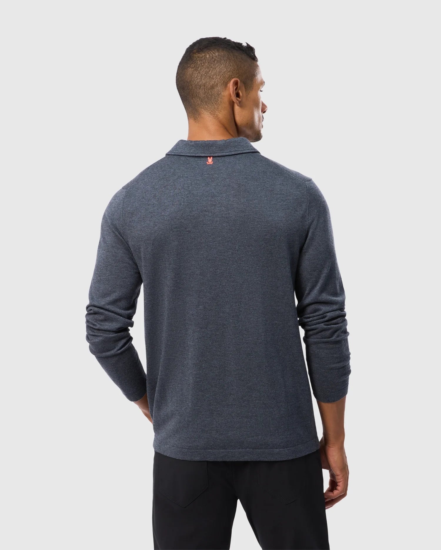 A man is turned away from the camera, wearing a long-sleeved MENS BRADSHAW POLO SWEATER - B6E835D200 in dark blue by Psycho Bunny and black pants. He is standing against a plain light gray background.