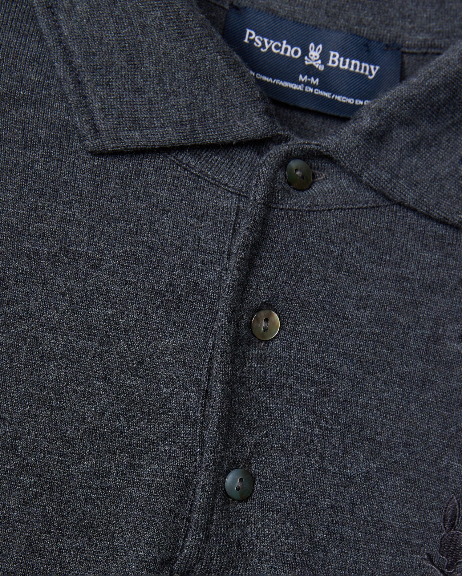 A close-up of the dark gray Men's Bradshaw Polo Sweater (B6E835D200) showcases its merino wool fabric, complete with buttons and a collar. The Psycho Bunny brand label is prominently displayed at the neck, featuring the iconic bunny logo that accentuates its luxurious appeal.