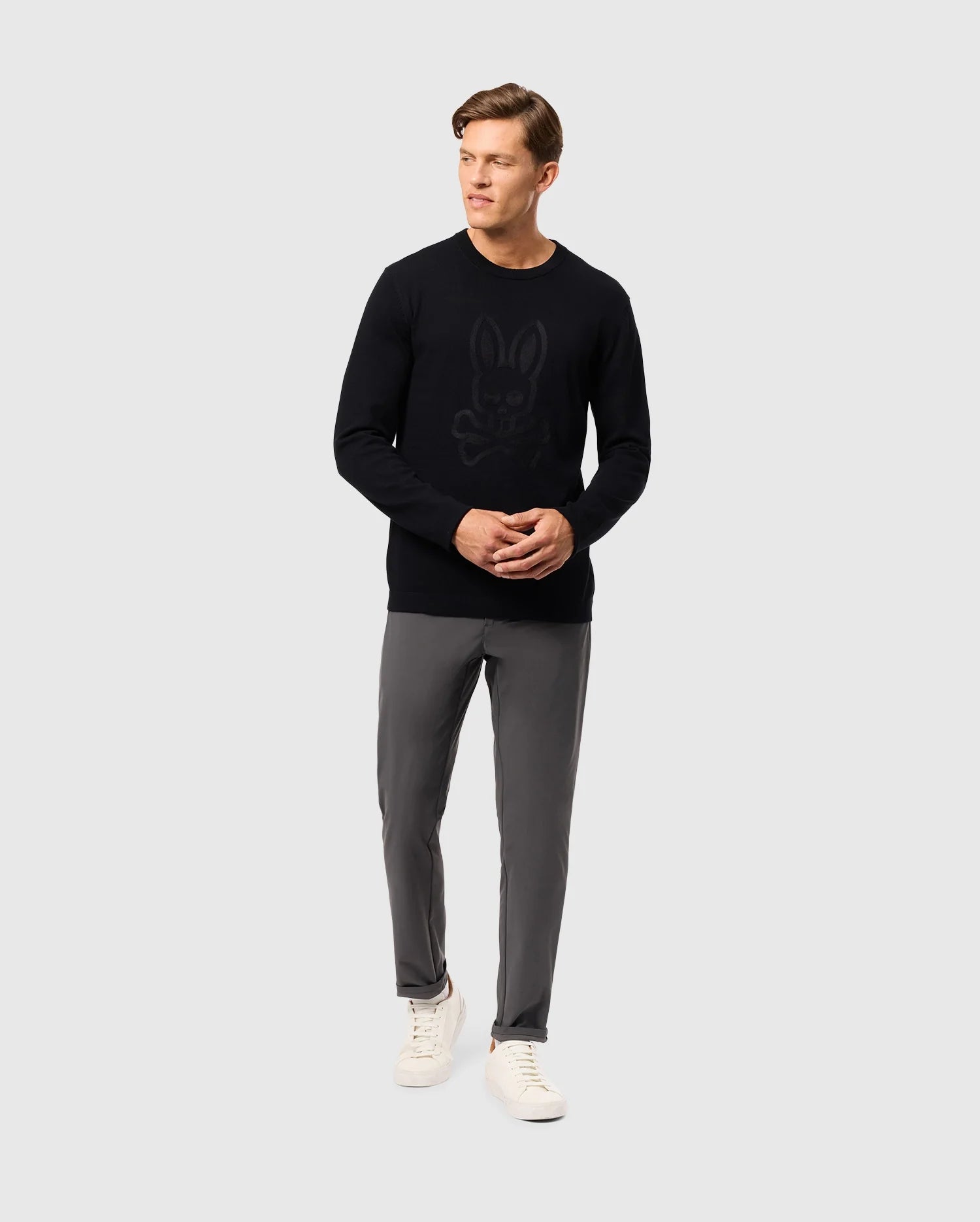 A man dressed in the Psycho Bunny MENS CORY CHAINSTITCH COTTON SWEATER, which is black and adorned with a subtle bunny design in chainstitch embroidery, pairs this stylish piece with gray pants and white sneakers while standing against a plain gray background.