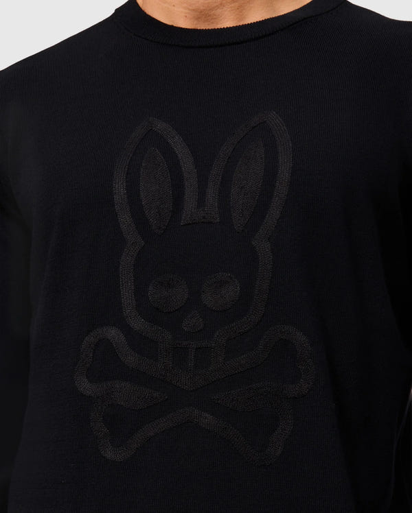 A person wearing a black Psycho Bunny Men’s Cory Chainstitch Cotton Sweater, featuring subtle chainstitch embroidery of a stylized bunny skull and crossbones on the front. The background is plain and light-colored.
