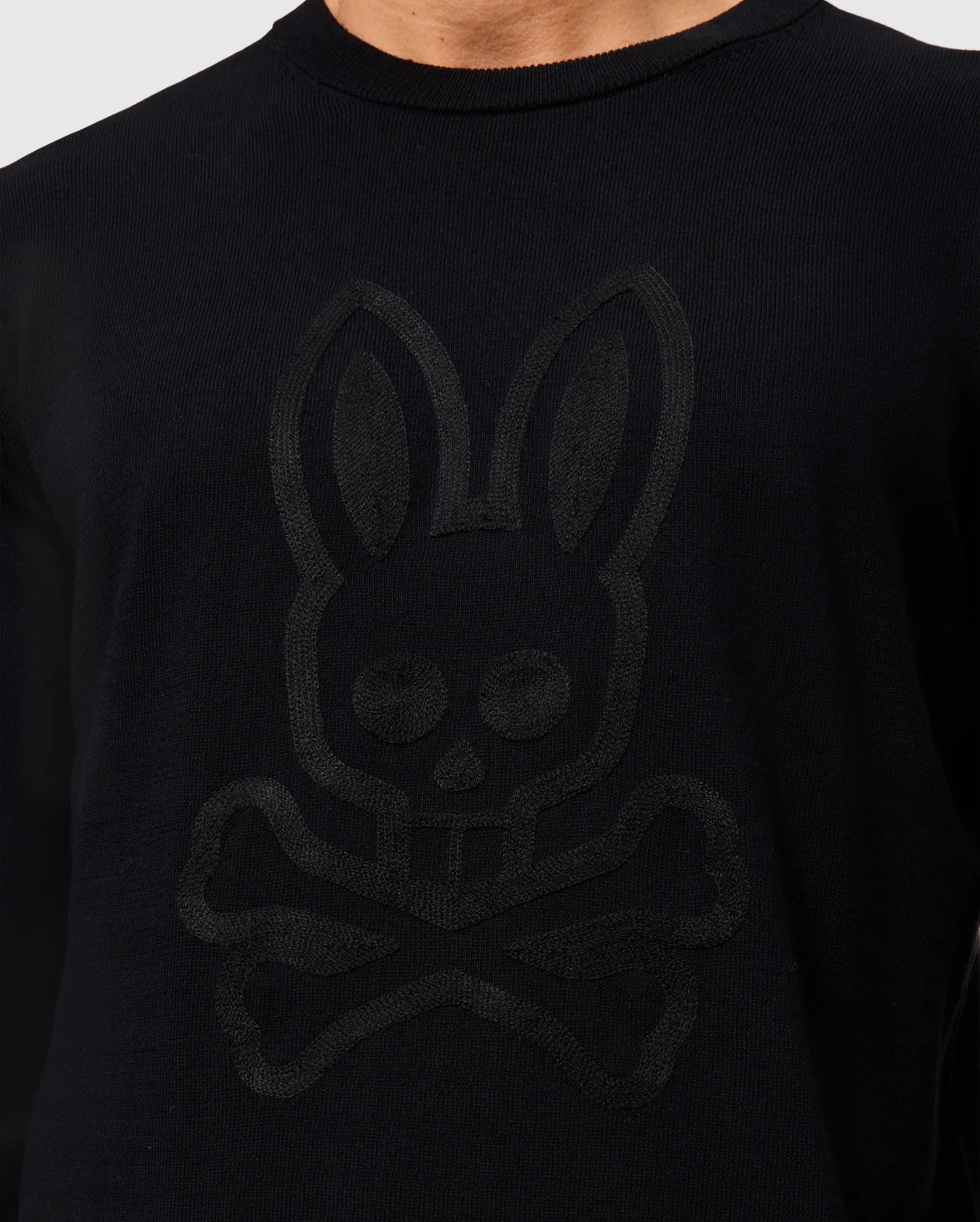 A person wearing a black Psycho Bunny Men’s Cory Chainstitch Cotton Sweater, featuring subtle chainstitch embroidery of a stylized bunny skull and crossbones on the front. The background is plain and light-colored.