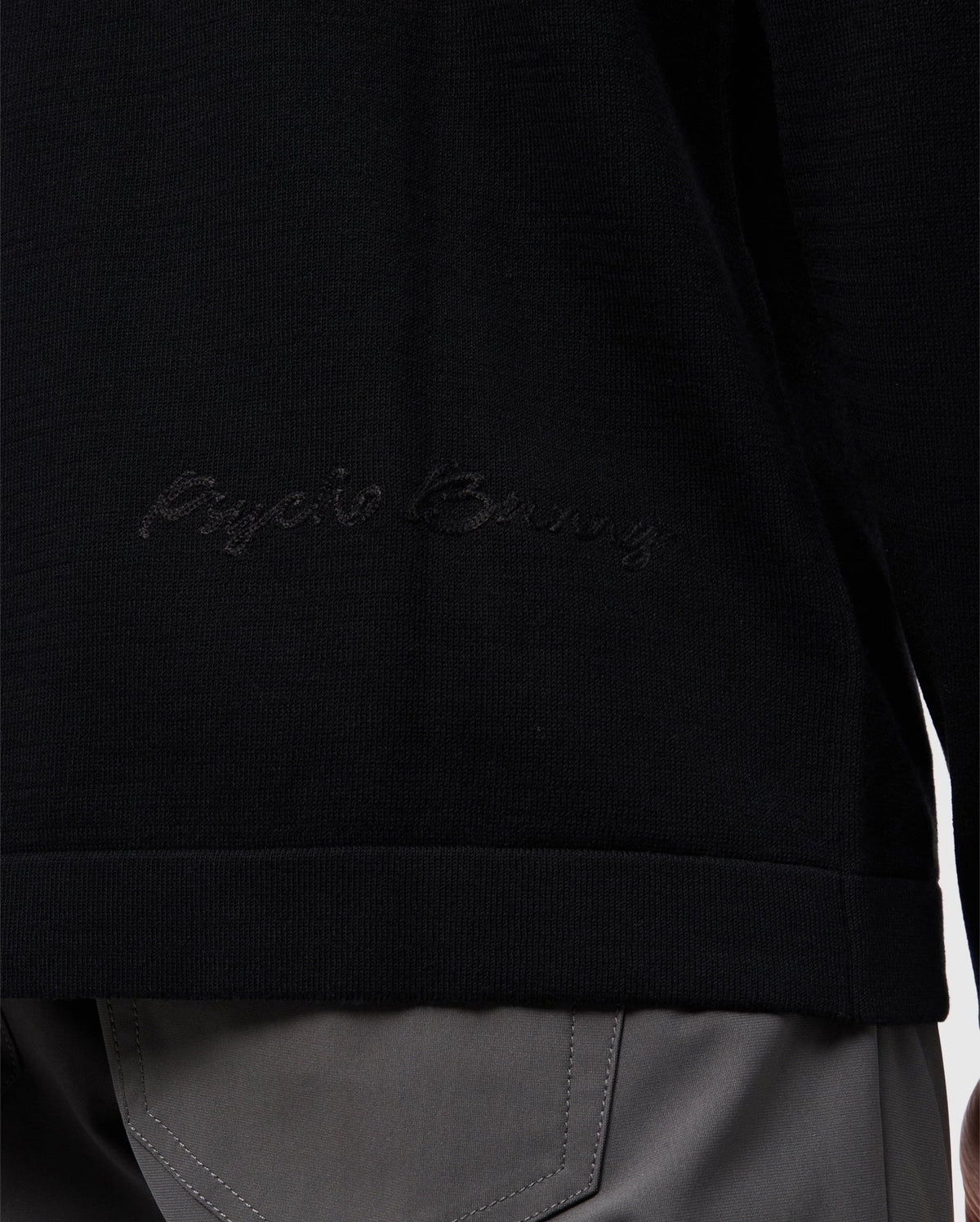 Close-up of a person wearing the Psycho Bunny MENS CORY CHAINSTITCH COTTON SWEATER - B6E893D200 in black, showcasing subtle chainstitch embroidery on the side. The lower half of the sweater is visible, paired with gray pants that have prominent stitching on the pockets.