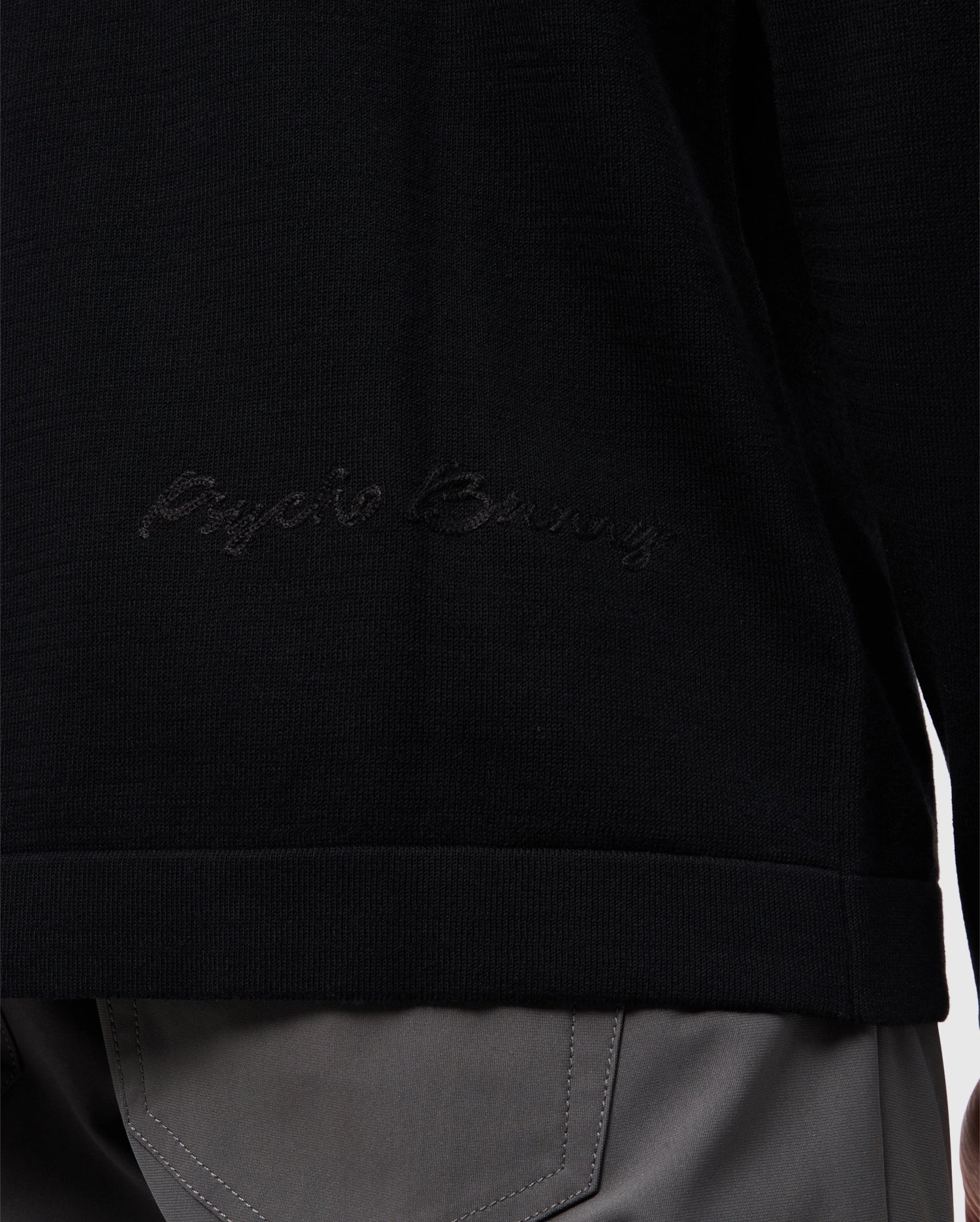 Close-up of a person wearing the Psycho Bunny MENS CORY CHAINSTITCH COTTON SWEATER - B6E893D200 in black, showcasing subtle chainstitch embroidery on the side. The lower half of the sweater is visible, paired with gray pants that have prominent stitching on the pockets.