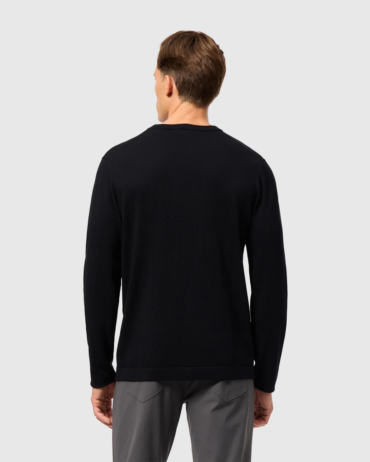 A person wearing the Psycho Bunny MENS CORY CHAINSTITCH COTTON SWEATER, a black long-sleeve garment with intricate chainstitch embroidery, paired with gray pants is facing away from the camera against a plain, light gray background.