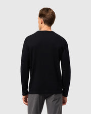 A person wearing the Psycho Bunny MENS CORY CHAINSTITCH COTTON SWEATER, a black long-sleeve garment with intricate chainstitch embroidery, paired with gray pants is facing away from the camera against a plain, light gray background.