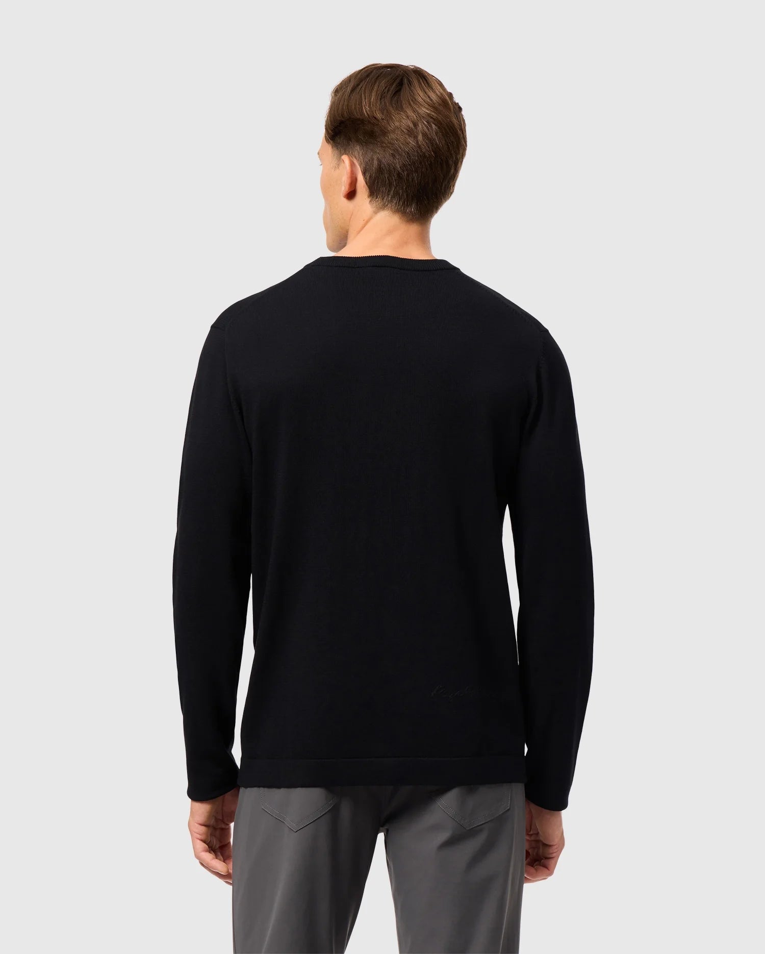 A person wearing the Psycho Bunny MENS CORY CHAINSTITCH COTTON SWEATER, a black long-sleeve garment with intricate chainstitch embroidery, paired with gray pants is facing away from the camera against a plain, light gray background.