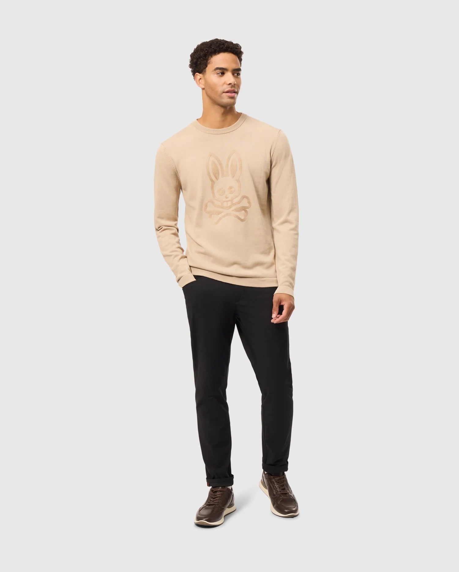 A person stands against a plain background, donning the Psycho Bunny MENS CORY CHAINSTITCH COTTON SWEATER - B6E893D200, which showcases intricate chainstitch embroidery with a rabbit and crossbones design. They pair the lightweight cotton sweater with black pants and brown shoes, casually resting one hand in their pocket while maintaining a relaxed expression.