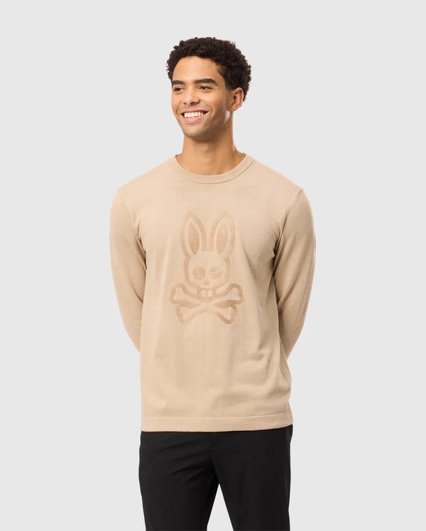 A smiling man wearing the Psycho Bunny MENS CORY CHAINSTITCH COTTON SWEATER - B6E893D200, featuring a bunny skull and crossbones chainstitch embroidery, stylishly pairs it with black pants against a light gray background.