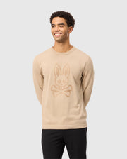 A smiling man wearing the Psycho Bunny MENS CORY CHAINSTITCH COTTON SWEATER - B6E893D200, featuring a bunny skull and crossbones chainstitch embroidery, stylishly pairs it with black pants against a light gray background.