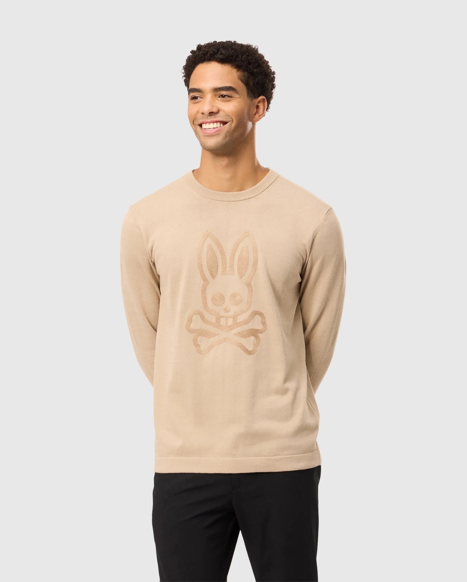 A smiling man wearing the Psycho Bunny MENS CORY CHAINSTITCH COTTON SWEATER - B6E893D200, featuring a bunny skull and crossbones chainstitch embroidery, stylishly pairs it with black pants against a light gray background.