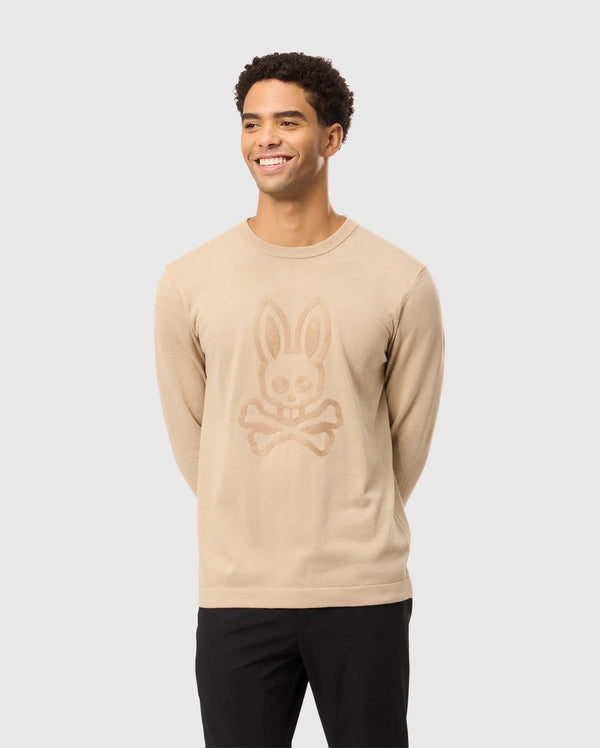 A smiling man wearing the Psycho Bunny MENS CORY CHAINSTITCH COTTON SWEATER - B6E893D200, featuring a bunny skull and crossbones chainstitch embroidery, stylishly pairs it with black pants against a light gray background.