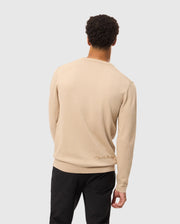 A person with short curly hair is seen from behind, dressed in a Psycho Bunny MENS CORY CHAINSTITCH COTTON SWEATER - B6E893D200 in beige and black pants. This lightweight cotton sweater showcases chainstitch embroidery with subtle text near the lower back. The background is plain and light gray.
