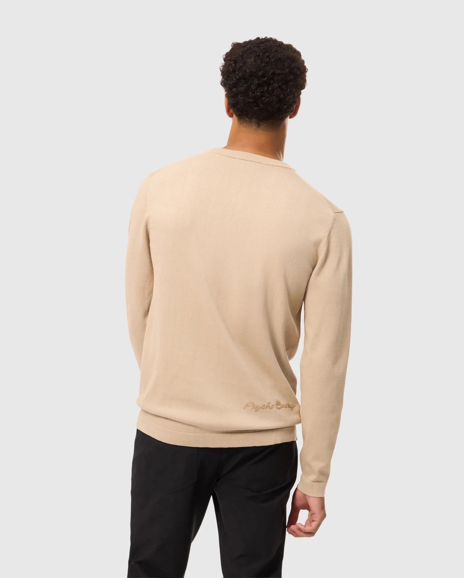 A person with short curly hair is seen from behind, dressed in a Psycho Bunny MENS CORY CHAINSTITCH COTTON SWEATER - B6E893D200 in beige and black pants. This lightweight cotton sweater showcases chainstitch embroidery with subtle text near the lower back. The background is plain and light gray.