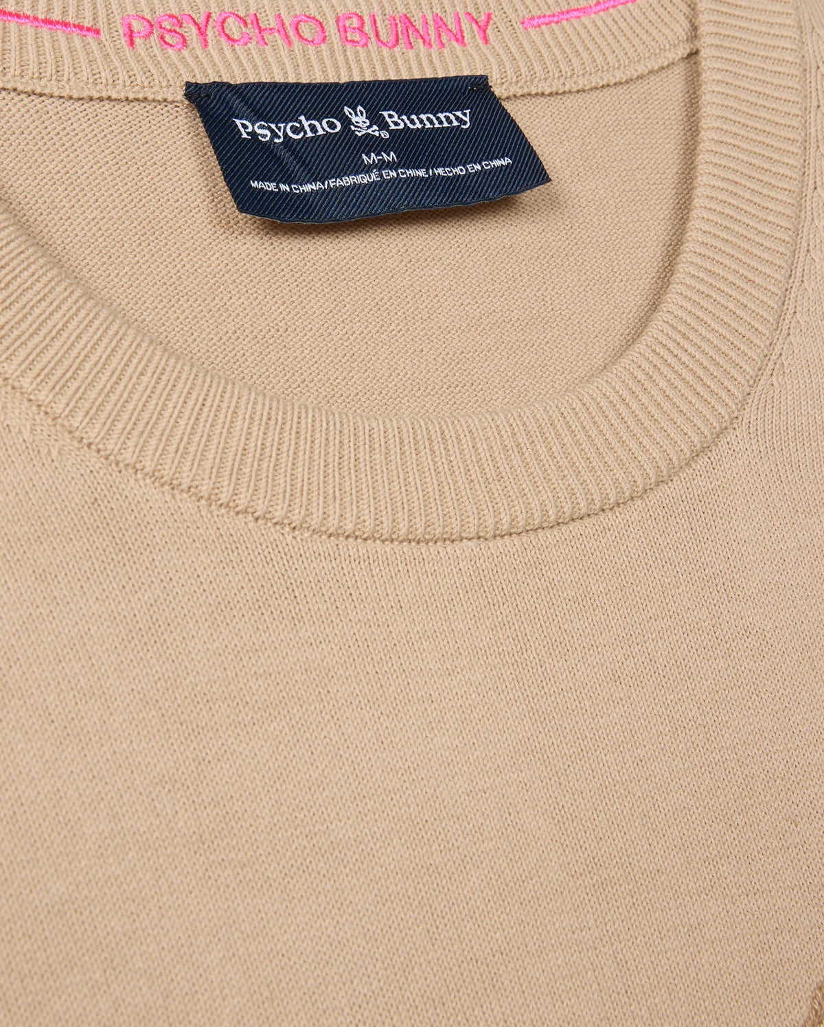 Close-up of a beige MENS CORY CHAINSTITCH COTTON SWEATER - B6E893D200 by Psycho Bunny, featuring a round neckline. The brand name "Psycho Bunny" appears in white on a dark tag, while "PSYCHO BUNNY" is crafted in pink chainstitch embroidery above the label, showcasing the expert craftsmanship of this lightweight cotton sweater.