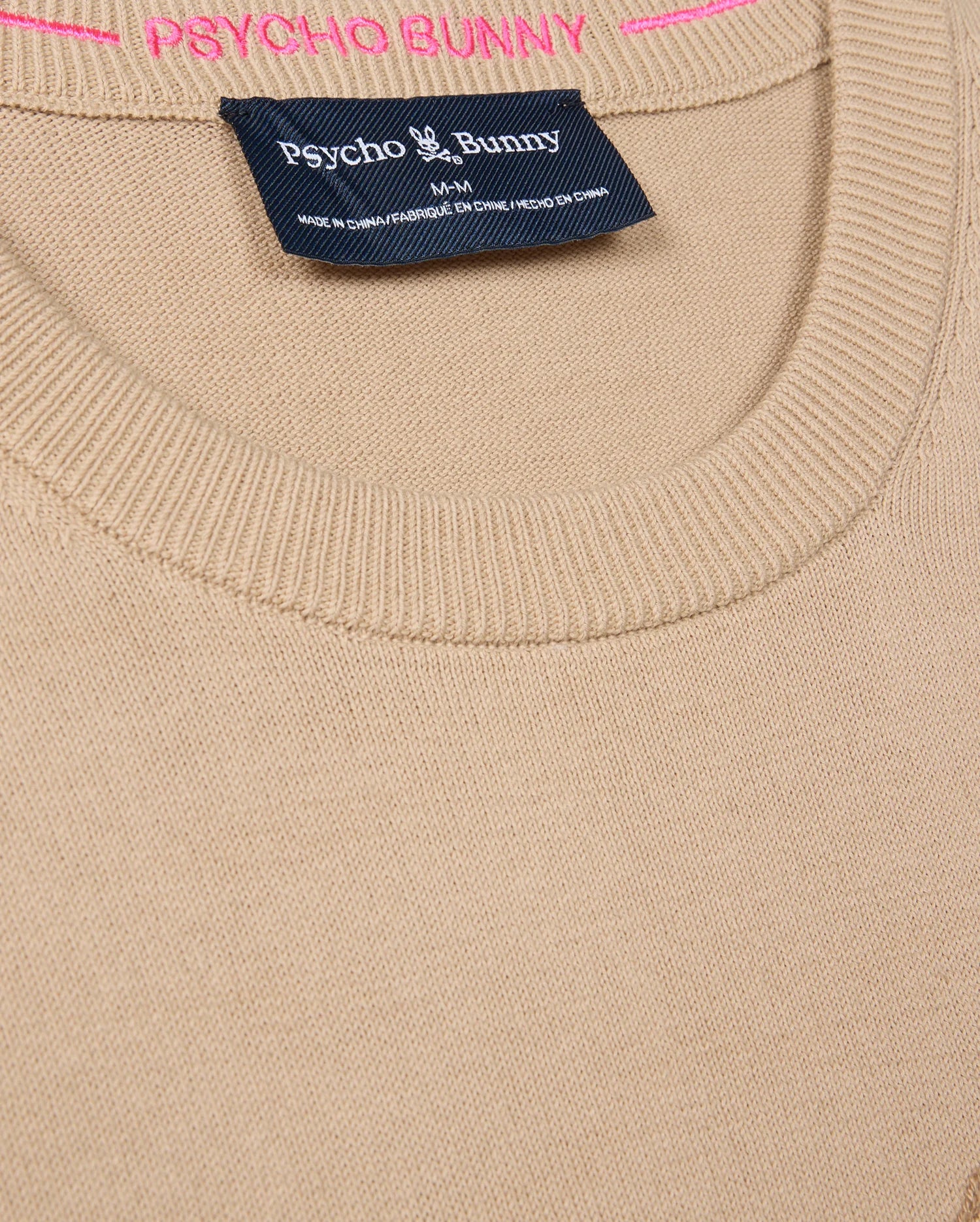 Close-up of a beige MENS CORY CHAINSTITCH COTTON SWEATER - B6E893D200 by Psycho Bunny, featuring a round neckline. The brand name "Psycho Bunny" appears in white on a dark tag, while "PSYCHO BUNNY" is crafted in pink chainstitch embroidery above the label, showcasing the expert craftsmanship of this lightweight cotton sweater.