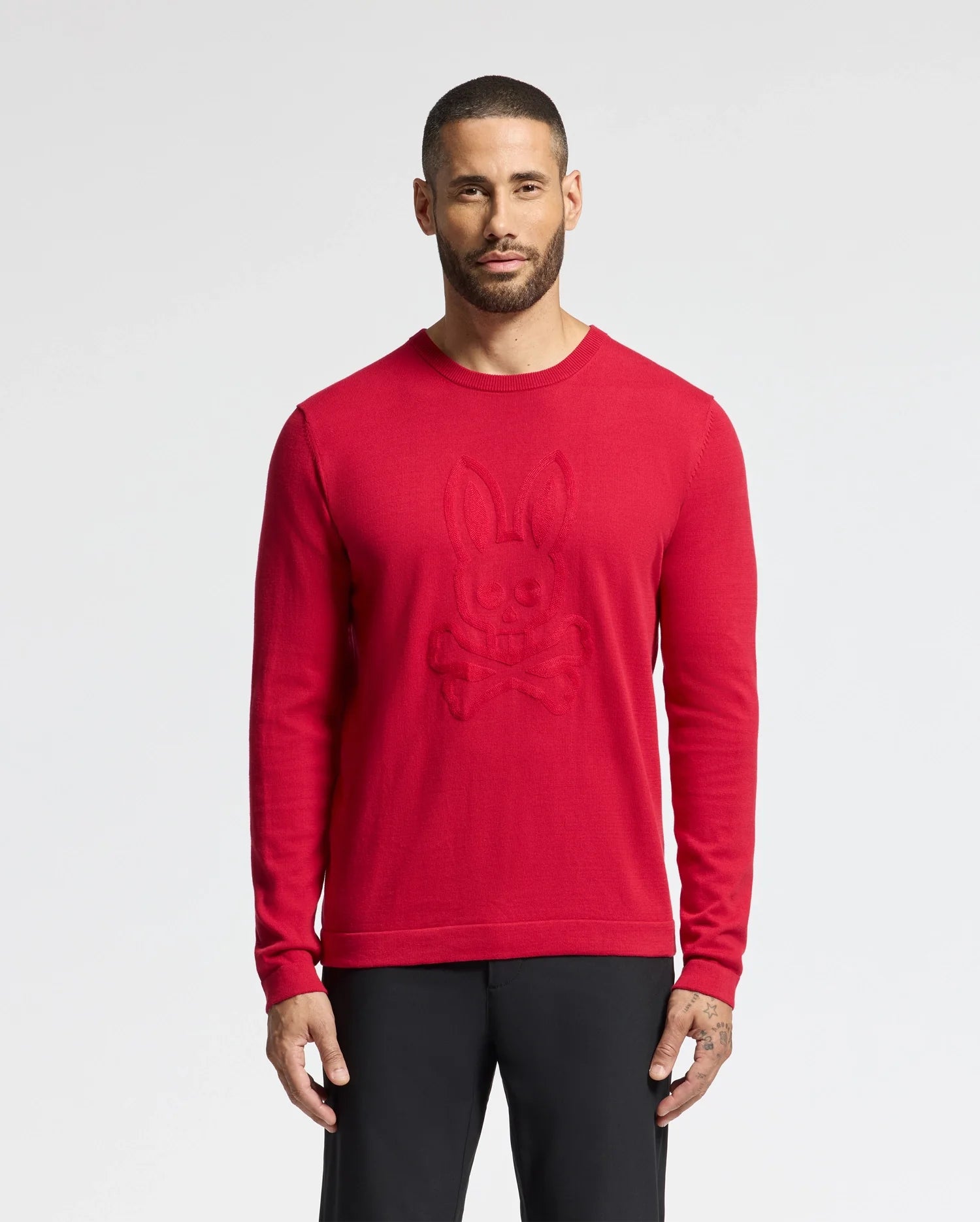 A short-haired, bearded person wears a Psycho Bunny MENS COORY CHAINSTITCH COTTON SWEATER featuring a vibrant skull and crossbones with bunny ears. The chainstitch embroidery adds texture. They're against a neutral background, paired with black pants.