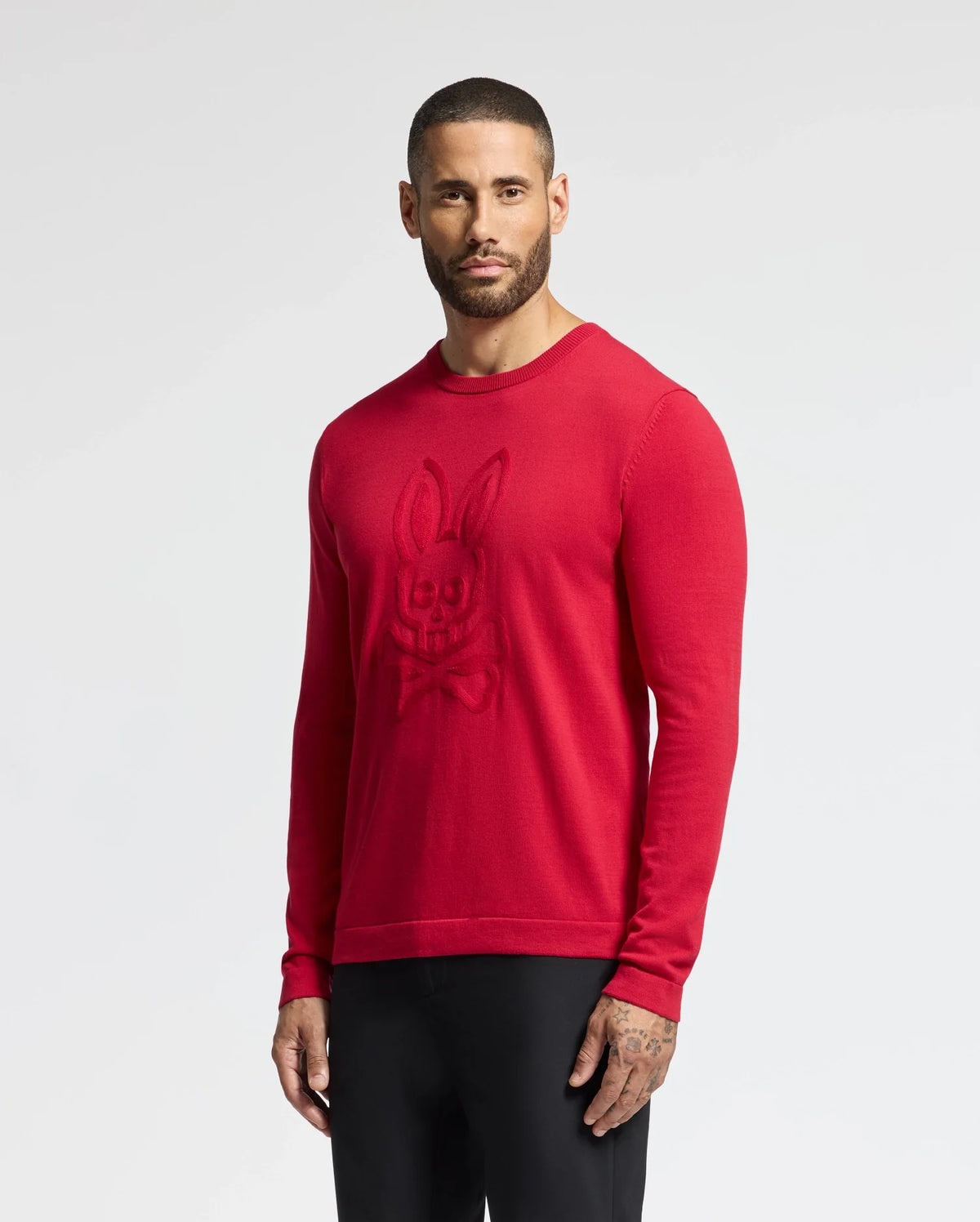 A man wears the MENS COORY CHAINSTITCH COTTON SWEATER by Psycho Bunny, featuring extra fine cotton and a chainstitch bunny logo, paired with black pants against a plain white background.