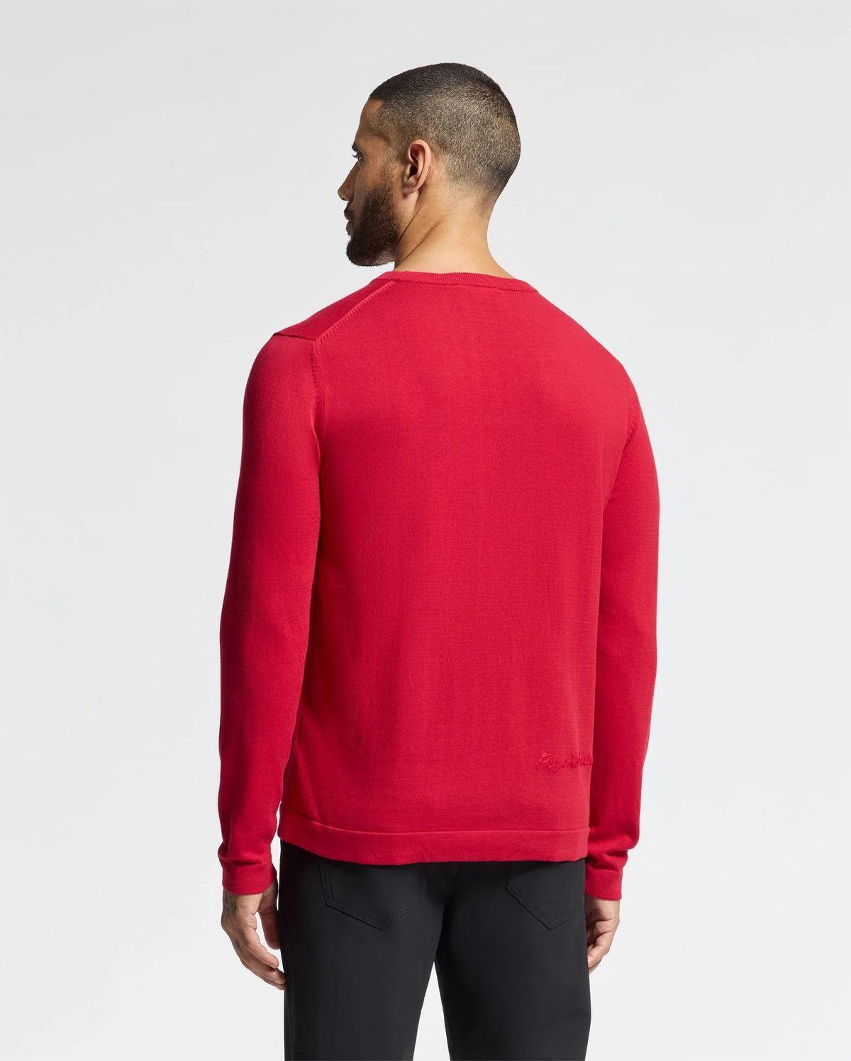 A person with short hair stands facing away from the camera wearing Psycho Bunny's MENS COORY CHAINSTITCH COTTON SWEATER - B6E893E200 in red and black pants. The long sleeves subtly highlight the chainstitch embroidery against a plain white background.