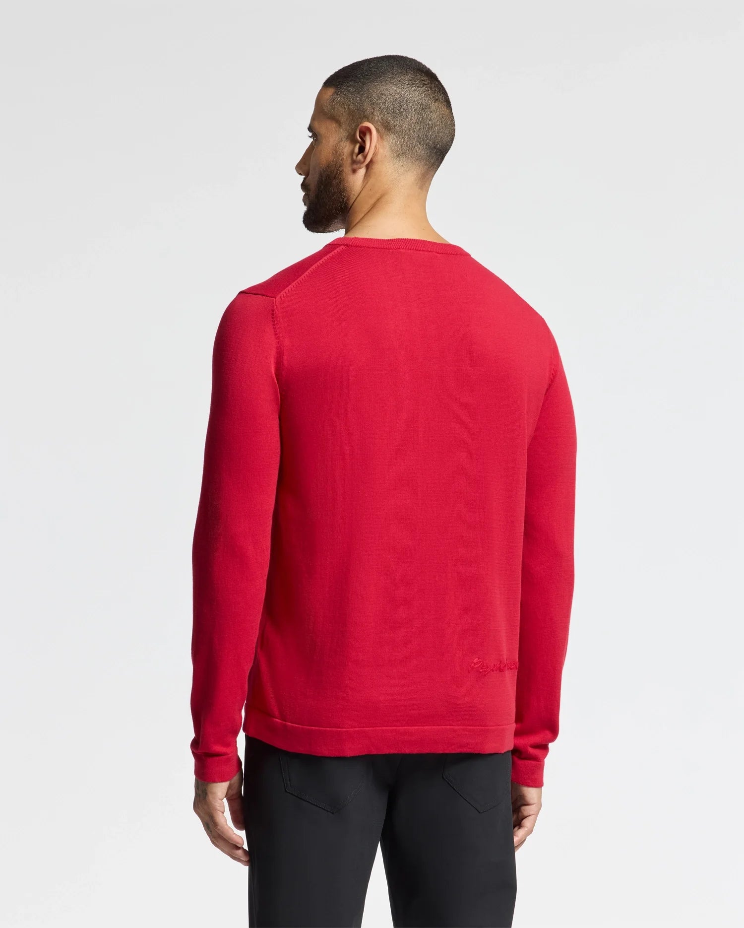 A person with short hair stands facing away from the camera wearing Psycho Bunny's MENS COORY CHAINSTITCH COTTON SWEATER - B6E893E200 in red and black pants. The long sleeves subtly highlight the chainstitch embroidery against a plain white background.
