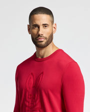 A bearded man wearing a red Psycho Bunny MENS COORY CHAINSTITCH COTTON SWEATER with a chainstitch bunny design stares at the camera against a plain backdrop.